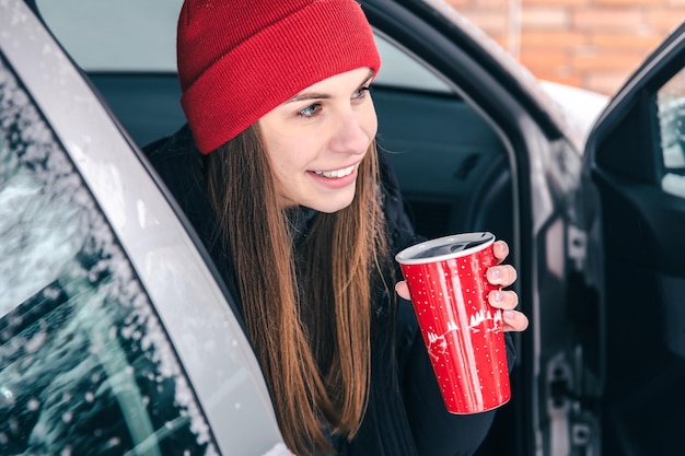 Glückliche junge Frau mit rotem Thermobecher sitzt im Winter in einem Auto