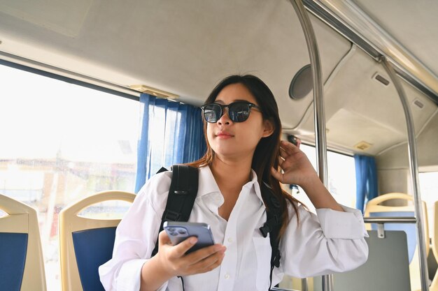 Glückliche junge Frau mit Sonnenbrille sitzt in einem öffentlichen Bus und genießt eine Reise.