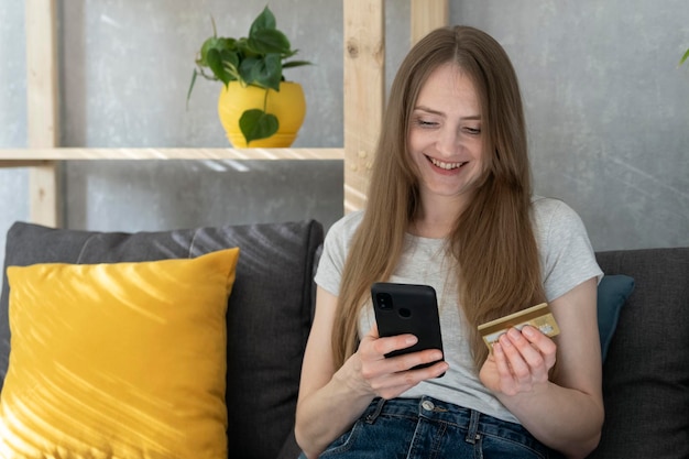 Glückliche junge Frau mit Telefon und Kreditkarte auf der Couch sitzen. Online Einkaufen.