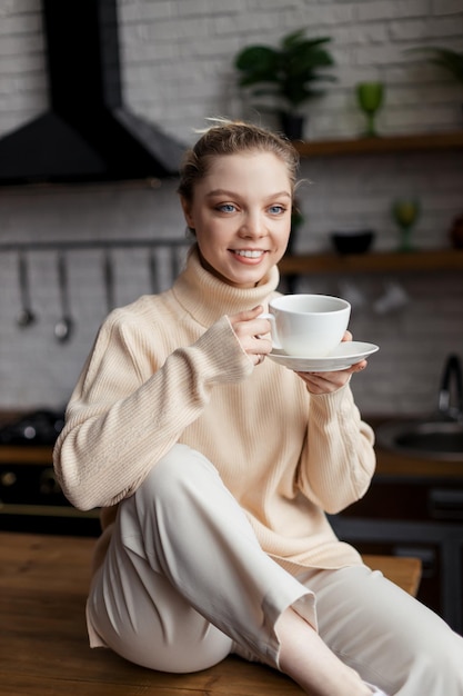 Glückliche junge Frau sitzt in gemütlicher Atmosphäre am Küchentisch und hält eine Tasse Kaffee