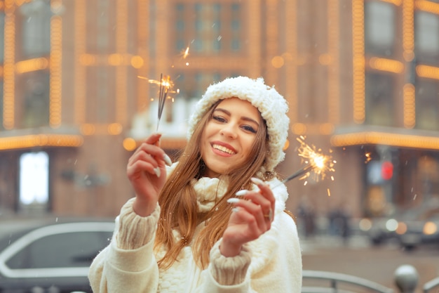Glückliche junge Frau trägt weiße Strickmütze und Mantel und hat Spaß mit Wunderkerzen auf der zu Weihnachten dekorierten Straße