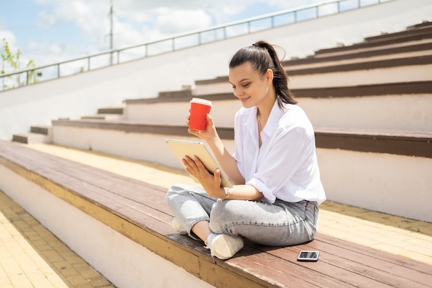 Glückliche junge Frauen mit Tablette, die Kaffee-Pappbecher hält und sonnigen Tag im Amphitheater genießt