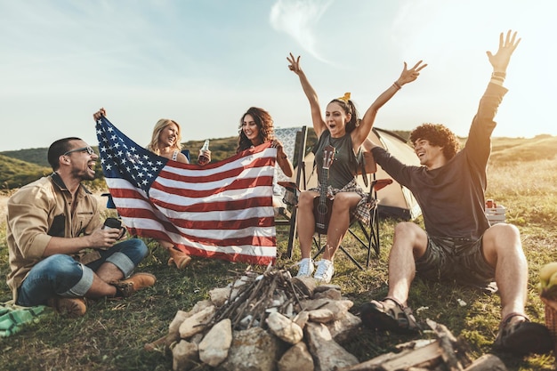 Glückliche junge Freunde feiern den 4. Juli und genießen einen sonnigen Tag am Berg. Sie halten die amerikanische Flagge, lachen und stoßen mit Bierflaschen an.