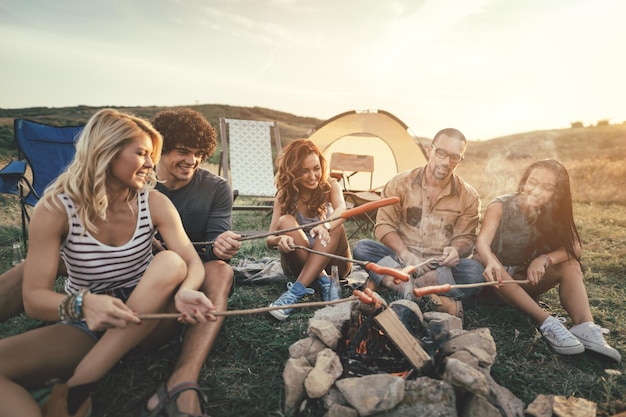 Glückliche junge Freunde genießen einen sonnigen Tag am Berg. Sie lachen und grillen Würste auf Stöcken über einem Lagerfeuer in der Nähe des Zeltes.