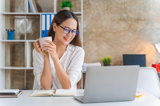 Glückliche junge Geschäftsfrau mit Laptop-Computer beim Online-Shopping Lächelnde schöne Asiatin, die am Arbeitsplatz sitzt und arbeitet