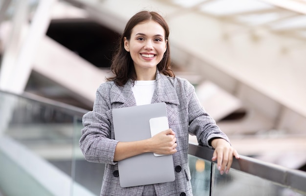 Glückliche junge Geschäftsfrau mit Laptop und Smartphone am modernen Flughafen