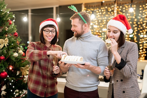 Glückliche junge Geschäftsfrauen in den Weihnachtsmannmützen, die Nachtisch und Champagner an der Weihnachtsfeier im Büro genießen