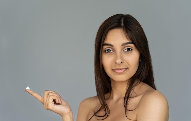 Glückliche junge indische Frau hält cremefarbenen Blick in die Kamera isoliert auf grauem Hintergrund