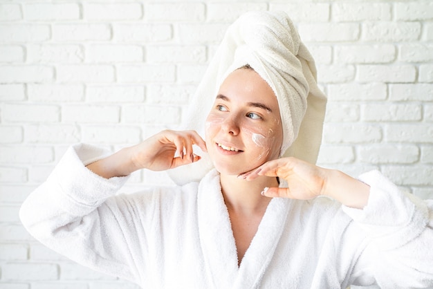 Glückliche junge kaukasische Frau im weißen Badetuch, das Gesichtscreme zu Hause anwendet