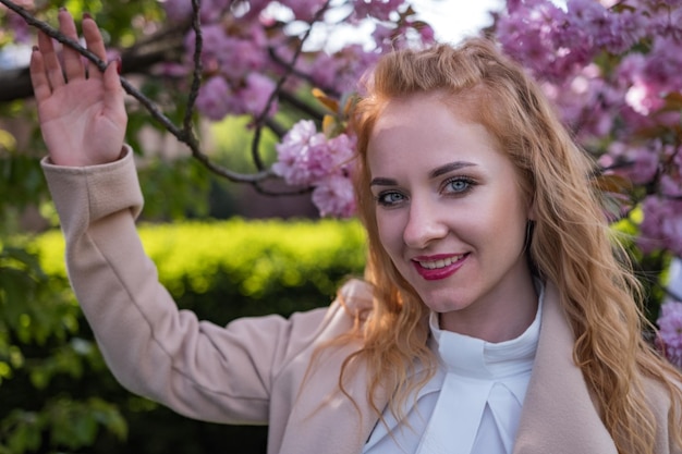 Glückliche junge kaukasische Frau mit rotem, welligem Haar auf blühendem Baumhintergrund Porträt eines charmanten, modischen Mädchens im blühenden Sakura-Park