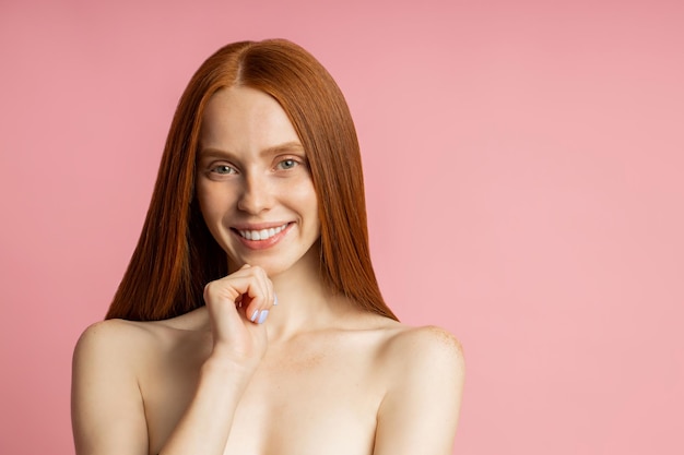 Glückliche junge kaukasische nackte Ingwerfrau mit sommersprossiger, sauberer Haut, die das Kinn berührt, lächelt zufrieden mit dem Ergebnis der Pflegeverfahren, Haarbehandlung, die über rosa Studiowand posiert.