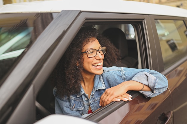 Glückliche junge lächelnde afroamerikanische Frau schwarzhaariger Fahrer mit Brille, die in einem neuen braunen Auto sitzt