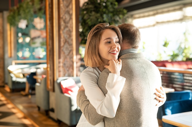 Glückliche junge lächelnde Frau, die ihren liebevollen Freund umarmt, während beide im luxuriösen Restaurant Liebe zueinander ausdrücken