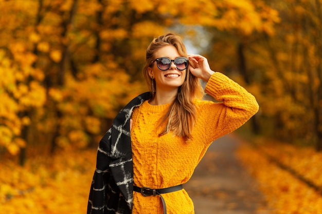 Glückliche junge lächelnde Frau mit stilvoller Sonnenbrille in modischem schwarzem Mantel und gestricktem gelbem Pullover geht und genießt im bunten Park mit Herbstblättern. Lustiges kaukasisches weibliches Model