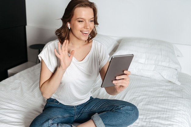 Glückliche junge lässig gekleidete Frau, die zu Hause auf dem Bett sitzt, Tablet-Computer hält und einen Chat hat