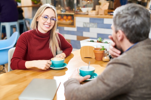 Glückliche junge lässige Geschäftsfrau, die Kaffee am Tisch im Café trinkt, während sie mit Kollegin vor ihr spricht