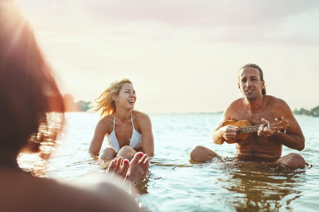 Glückliche junge Leute, die zusammen eine tolle Zeit am Strand haben. Sie sitzen im Wasser und ein Mann spielt Ukulele und singt. Sonnenuntergang über dem Wasser.