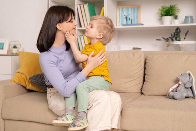 Glückliche junge liebevolle Mutter und ihr süßer kleiner Sohn, die Liebe miteinander ausdrücken, während sie auf weicher Couch im Wohnzimmer zu Hause sitzen