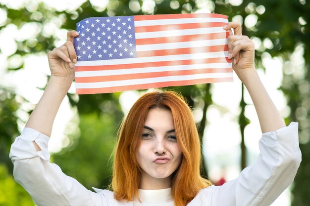 Glückliche junge lustige Frau, die mit der Nationalflagge der USA aufwirft, die es in ihren ausgestreckten Händen hält, die draußen im Sommerpark stehen. Positives Mädchen, das den Unabhängigkeitstag der Vereinigten Staaten feiert.