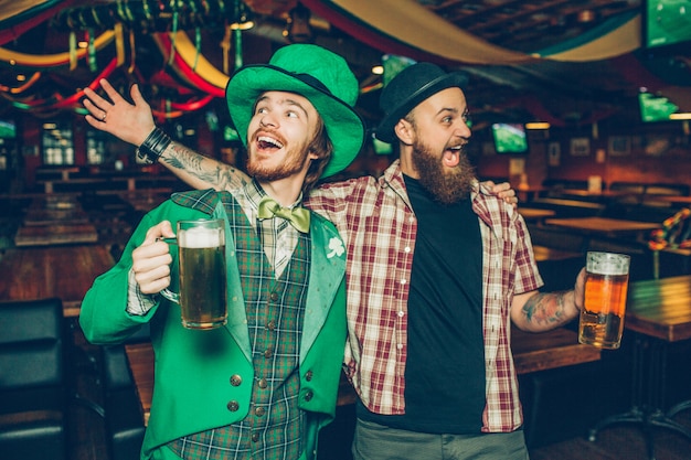 Glückliche junge Männer, die Becher Bier halten und zusammen in der Kneipe singen. Sie feiern den Saitn Patrick's Day. Guy auf der linken Seite tragen grünen Anzug.