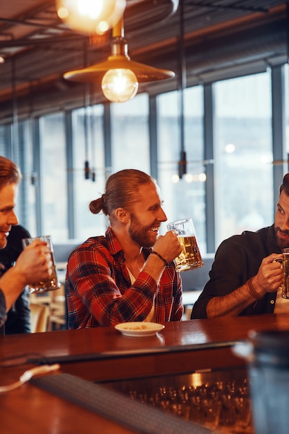 Glückliche junge Männer in Freizeitkleidung, die Bier genießen und lächeln, während sie an der Bartheke in der Kneipe sitzen