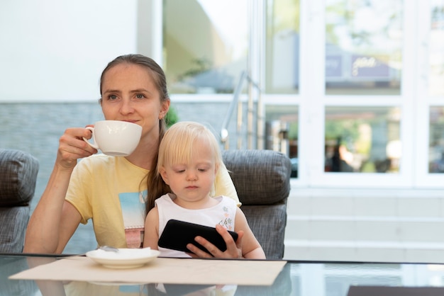 Glückliche junge Mutter trinkt Kaffee und hält Kind in ihren Armen. Kind beobachtet Cartoons am Telefon, während Mama entspannt ist.