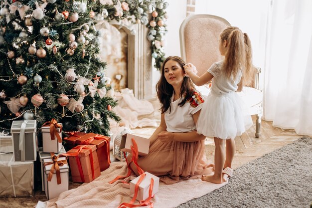 Glückliche junge Mutter und ihre kleine Tochter in schönem Kleid sitzen in der Nähe des Neujahrsbaums und öffnen Neujahrsgeschenke im hellen, gemütlichen Zimmer.