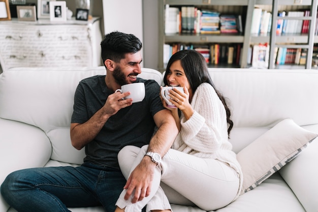 Glückliche junge Paare, die auf dem Sofa hält weiße Kaffeetasse sitzen