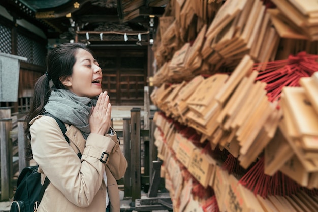 glückliche junge reisende, die mit traditionellem hölzernem gebetsbrett am tenmangu-schrein in osaka betet. Fröhliches Touristenmädchen schließt die Augen und legt Palmen zusammen, die an der Wunschwand im Tempel im Freien stehen