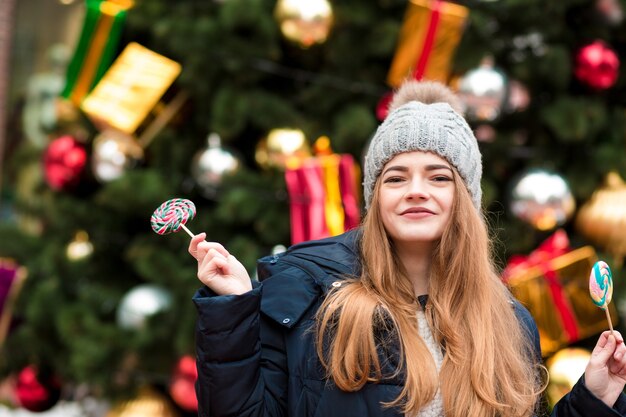 Glückliche junge rothaarige Frau in grauer Strickmütze, die köstliche Karamellbonbons gegen Weihnachtsfichte hält