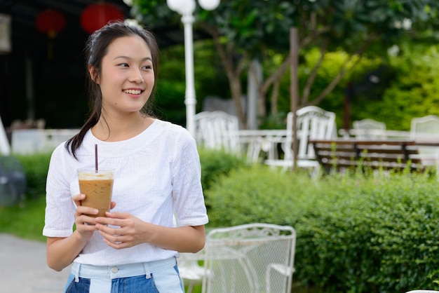 Glückliche junge schöne asiatische Frau, die am Kaffeehaus draußen denkt