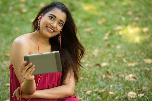 Glückliche junge schöne indische Frau, die beim Verwenden des digitalen Tabletts am Park denkt