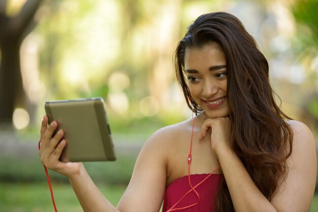 Glückliche junge schöne indische Frau, die digitales Tablett am Park verwendet