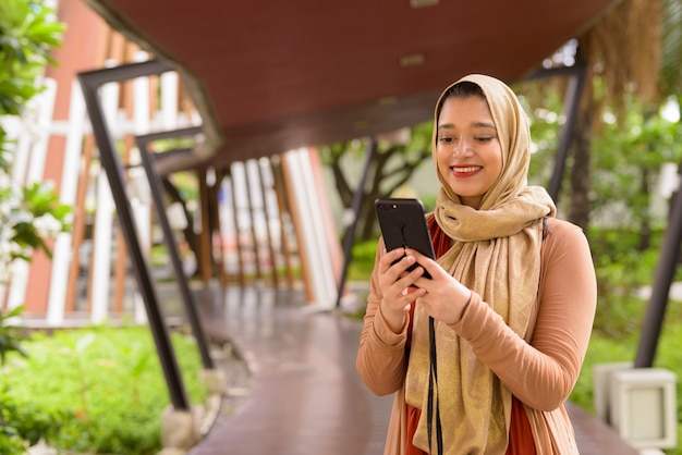 Glückliche junge schöne indische muslimische Frau, die Telefon in der Stadt mit Natur verwendet