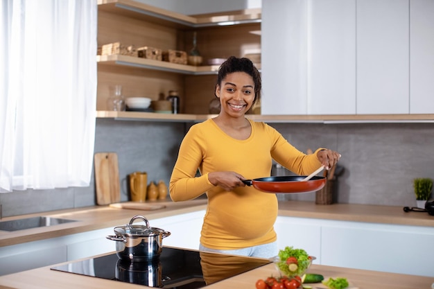 Glückliche junge schwangere Afroamerikanerin mit dickem Bauch, der Bratpfanne hält und Essen kocht