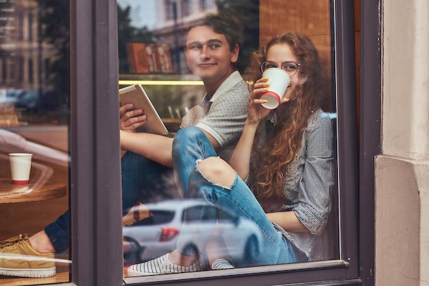 Glückliche junge Studenten, die während einer Pause Kaffee trinken und ein digitales Tablet auf einem Fensterbrett auf einem College-Campus verwenden.