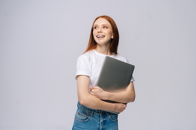 Glückliche junge Studentin, die Laptop-Computer hält und auf isoliertem grauem Hintergrund wegschaut