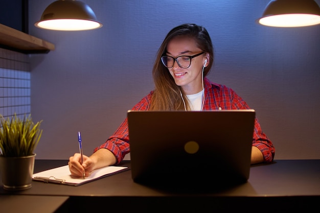 Glückliche junge Studentin in Brille und Kopfhörer, die Online-Webinar-Kurse sieht und per Konferenzvideoanruf kommuniziert. E Lernen und Prüfungsvorbereitung mit einem Laptop zu Hause