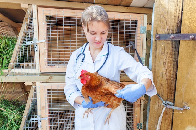 Glückliche junge tierarztfrau mit stethoskop, das huhn auf ranch hält und untersucht