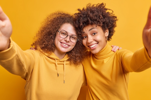 Foto glückliche junge, vielfältige frauen haben freundschaftliche beziehungen umarmen und strecken die arme nach vorne pose für selfie tragen freizeitkleidung lächeln angenehm isoliert über gelber wand haben spaß zusammen.