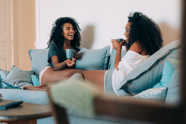 Glückliche junge zwei schwarze Frauen, die im trinkenden Kaffee der Couch sitzen