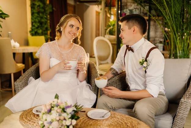 Glückliche Jungvermählten schauen sich an und trinken Kaffee, halten Tassen mit Kaffee in den Händen. Liebesparade in einem gemütlichen Interieur ein Liebespaar vor der Hochzeitszeremonie.