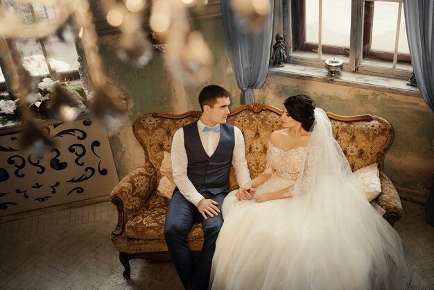 Glückliche Jungvermählten sitzen auf der Couch in einem Vintage-Interieur und schauen sich Händchen haltend an. Schönes Paar, Ehemann und Ehefrau, Braut und Bräutigam. Liebe, Hochzeitskonzept.