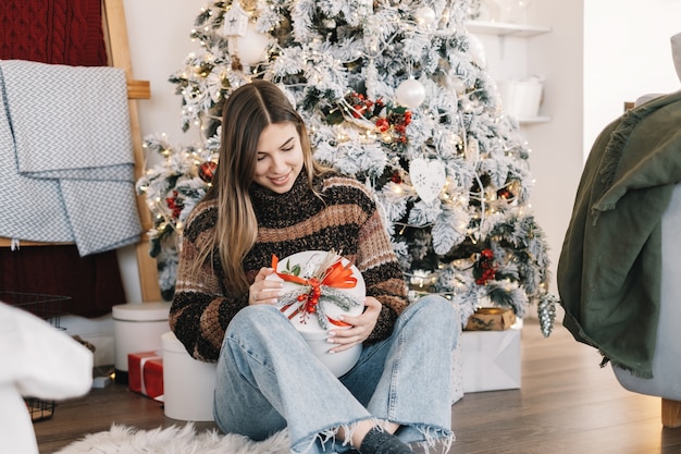 Glückliche kaukasische Frau, die großes Weihnachtsgeschenk hält, während auf dem Boden zu Hause nahe Weihnachtsbaum sitzt.