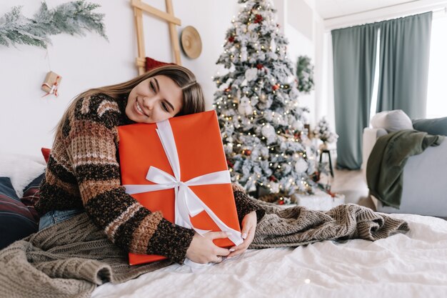 Glückliche kaukasische Frau, die großes Weihnachtsgeschenk hält, während auf einem Bett zu Hause nahe Weihnachtsbaum sitzt.