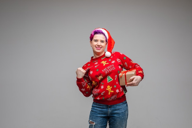 Foto glückliche kaukasische frau in der weihnachtsmütze und im wintertrikot und in den jeans, die ihre faust ballen und das eingewickelte weihnachtsgeschenk halten und lächeln