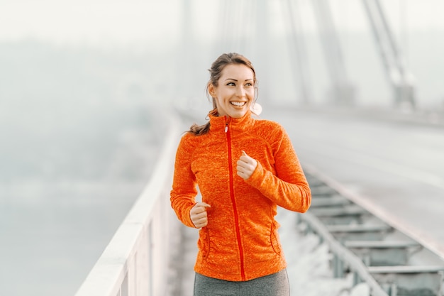 Glückliche kaukasische Frau in Sportbekleidung und mit Pferdeschwanz, der auf der Brücke im Winter läuft. Winter Fitness Konzept.