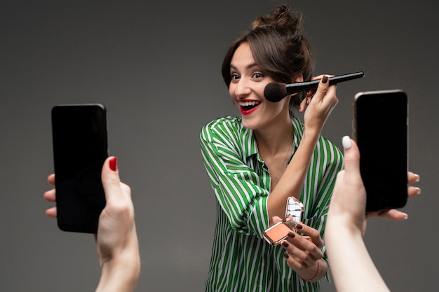 Glückliche kaukasische Frau Make-up mit Pinseln vor dem Spiegel und macht Fotos isoliert auf grauer Wand