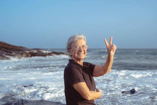 Glückliche kaukasische Seniorin, die bei Sonnenuntergang am Strand steht und die Kamera mit dem Daumen nach oben über dem Meer und blauem Himmel betrachtet