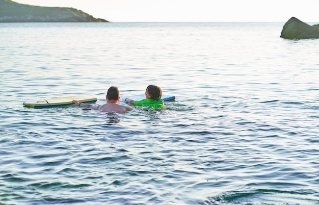 Glückliche Kinder, die die Kamera betrachten, die mit Bodyboard im Meer spielt. Kinder haben Spaß im Freien. Konzept der Sommerferien und des gesunden Lebensstils.